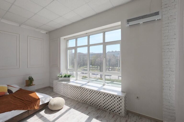Un open space bianco con la vetrata sul viale alberato, il letto basso e un pouf