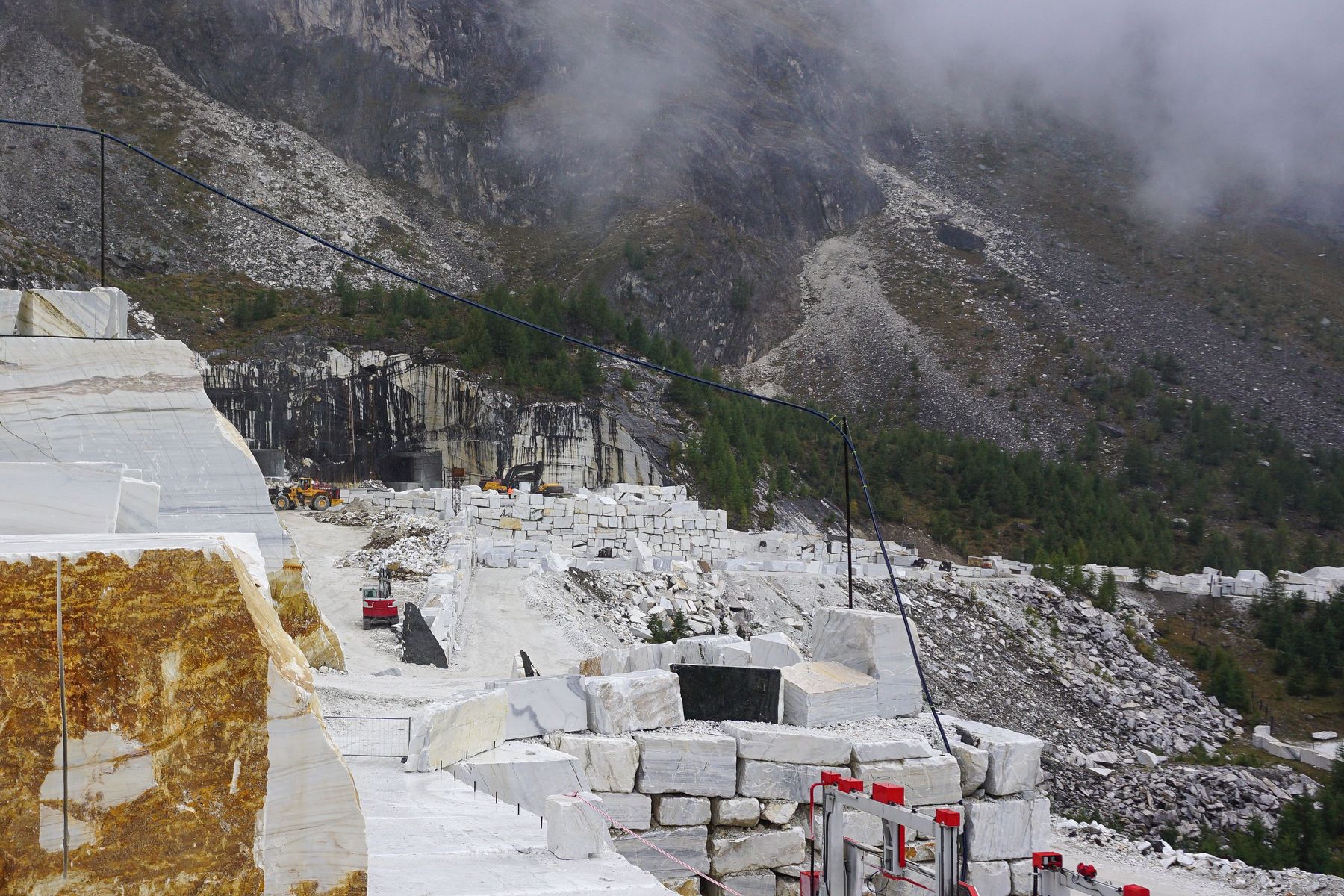 Il piazzale della cava coi blocchi bianchi squadrati e impilati, la tagliatrice a filo rossa e la montagna nella nebbia