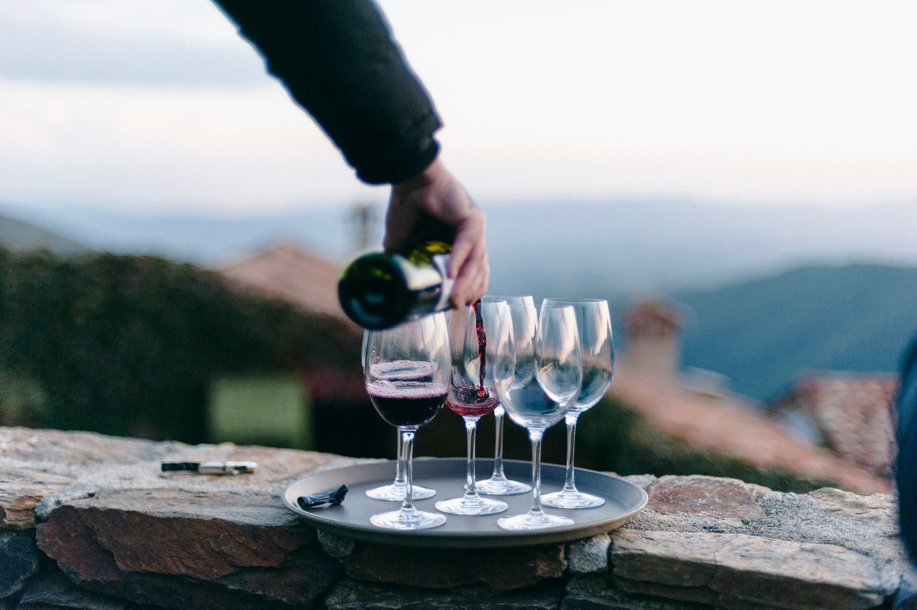 Vino rosso versato nei calici su un muretto di pietra, con le colline al tramonto