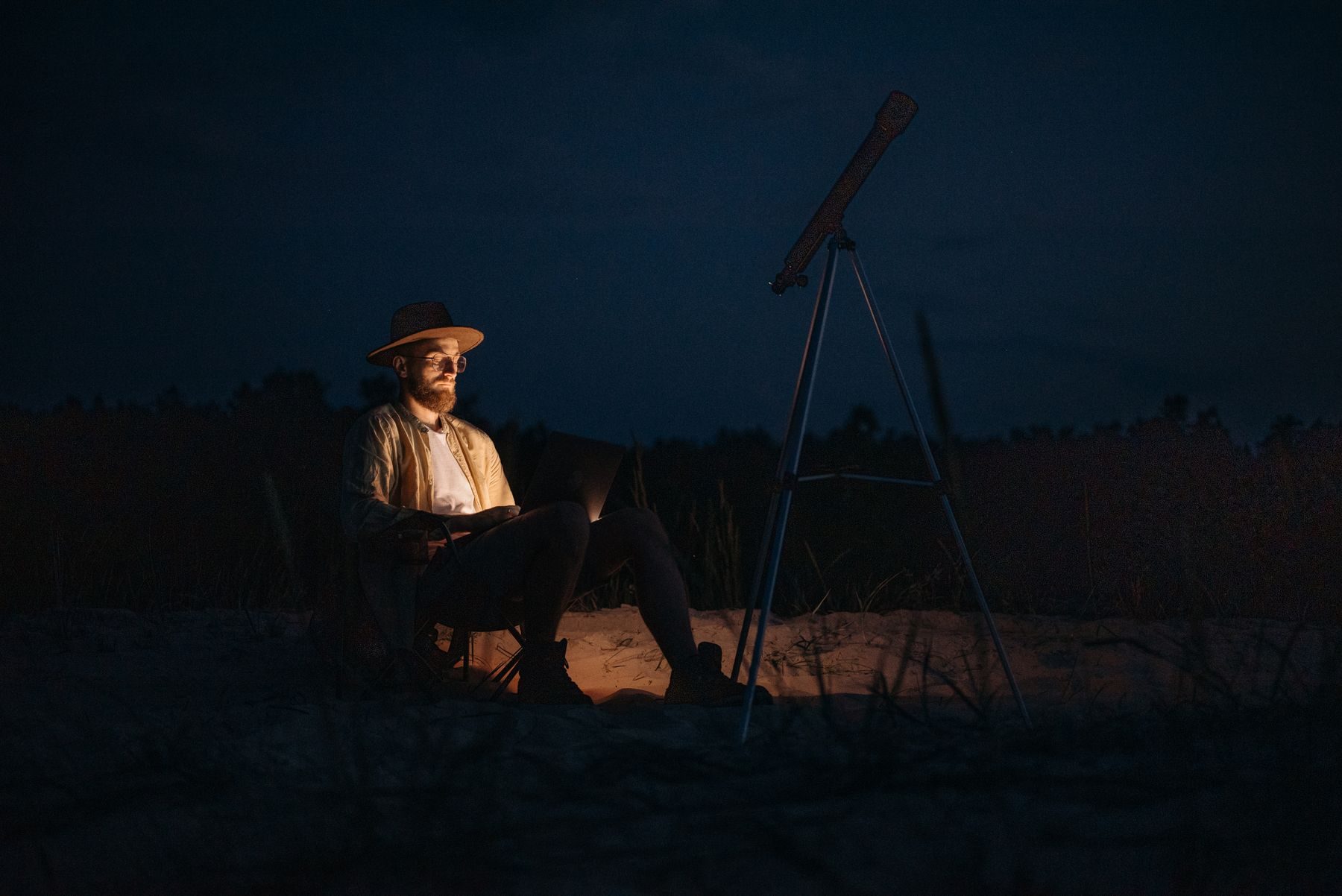 Un uomo col cappello e il portatile accanto al telescopio, di notte